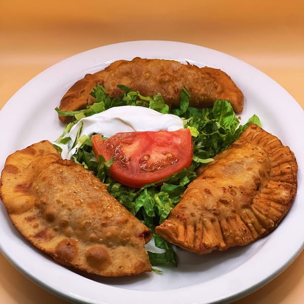 Three golden-brown empanadas arranged on a white plate with a side salad of lettuce, tomato, and sour cream.