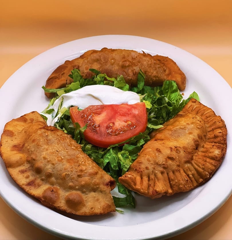 Three golden-brown empanadas arranged on a white plate with a side salad of lettuce, tomato, and sour cream.