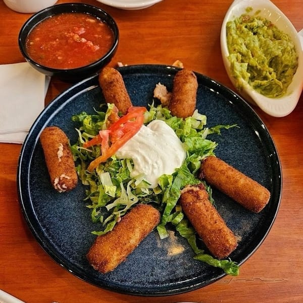 Fried taquitos with lettuce, sour cream, tomato, salsa, and guacamole on a wooden table.